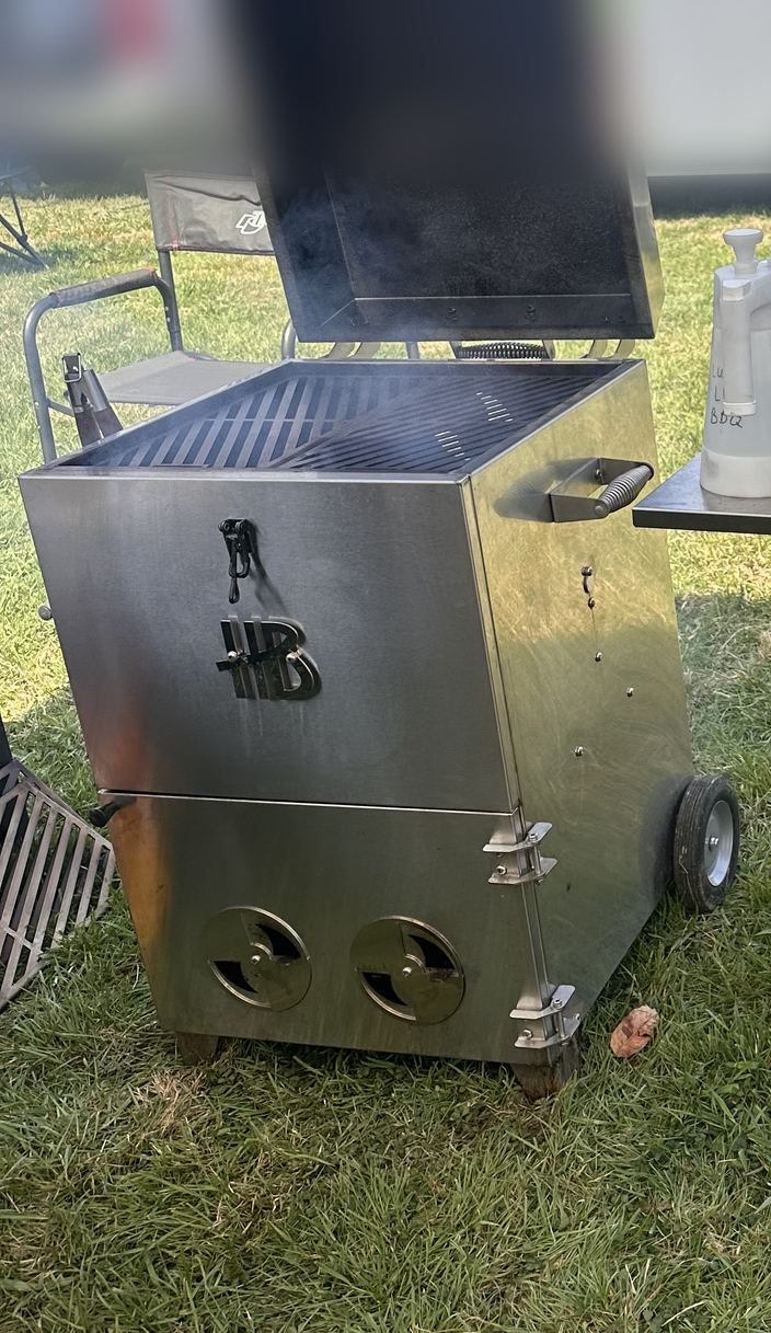The Hasty Bake Stainless Steel Roughneck, with the HB logo and two round vents, set up on the grass