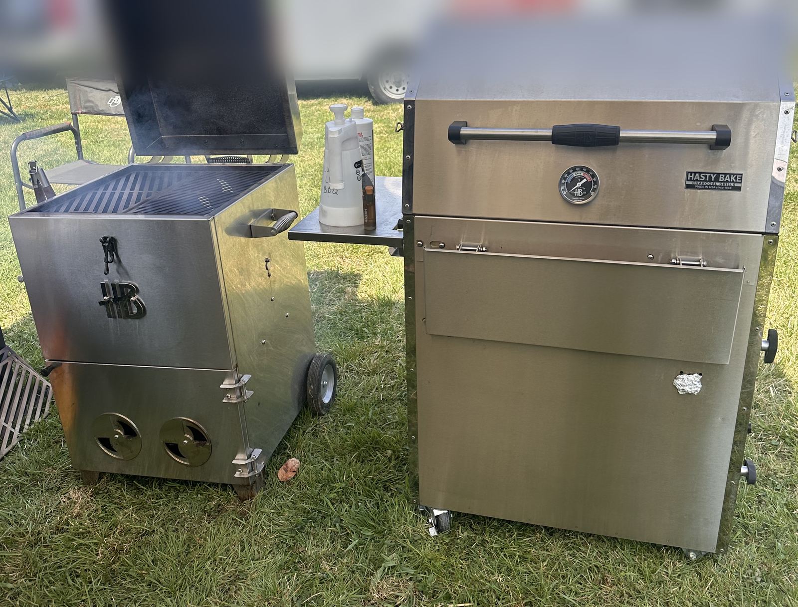 Two stainless Hasty Bake charcoal cookers set up on the grass at a cook site