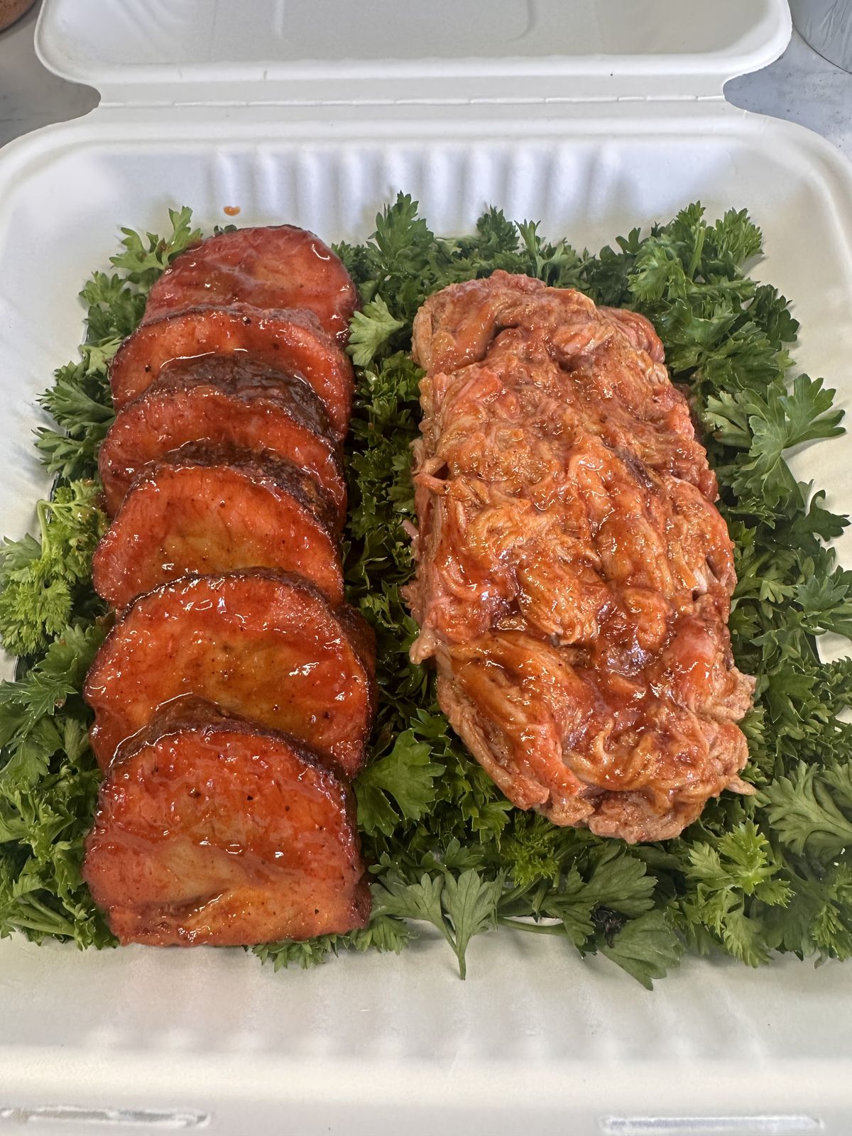 Takeout box of sliced, glazed pork tenderloin medallions next to sauced pulled pork on a bed of parsley